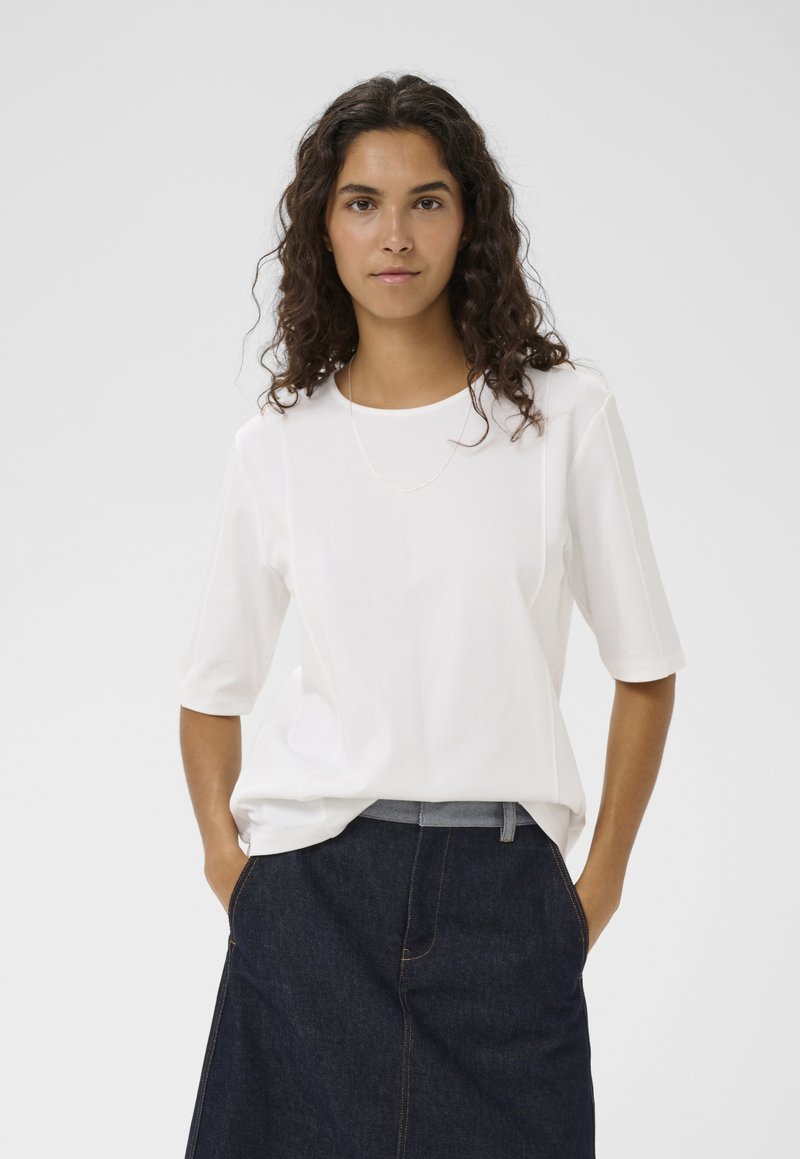 Femme aux cheveux bouclés et foncés, portant une chemise blanche à manches courtes et une jupe en jean foncé, debout les mains dans les poches devant un fond uni.