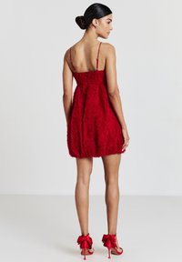 Frau in einem roten strukturierten ärmellosen Kleid mit dünnen Trägern und roten High Heels mit großen Schleifen, die mit dem Rücken zu einer einfarbigen Hintergrund steht.