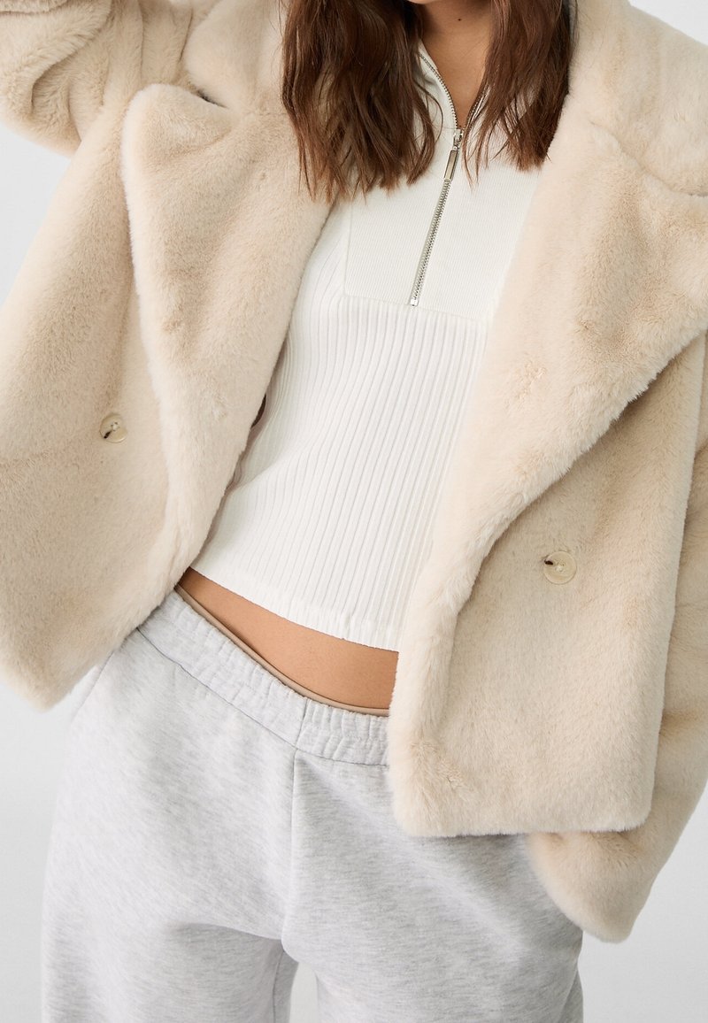 Beige Kunstpelzjacke mit weicher Textur, mit doppelreihigem Design, getragen über einem weißen, gerippten Top und grauen Jogginghosen.