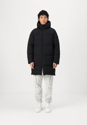 Black puffer coat with a hood, featuring a vertical quilted design and a front zipper. Paired with light gray cargo pants and white sneakers.