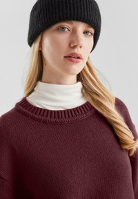Bordeaux gebreid truitje met een geribde halslijn gedragen over een witte coltrui, aangevuld met een zwarte geribde beanie. Soepele textuur en gelaagd ontwerp.