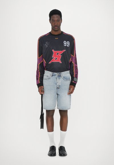 Sportliches schwarzes Langarmshirt mit grafischen Mustern, kombiniert mit hellblauen Jeansshorts, weißen Socken und schwarzen Slippern. Schwarze Tasche in der Hand.