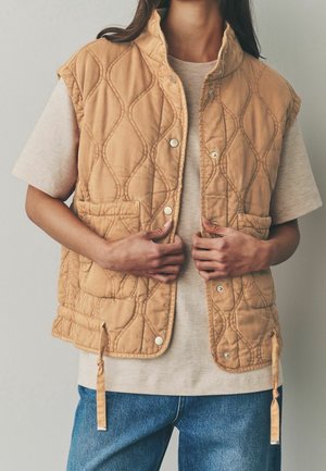 Person iført en beige quiltet vest over en beige kortærmet skjorte og blå jeans, justerer vestens forreste trykknapper.