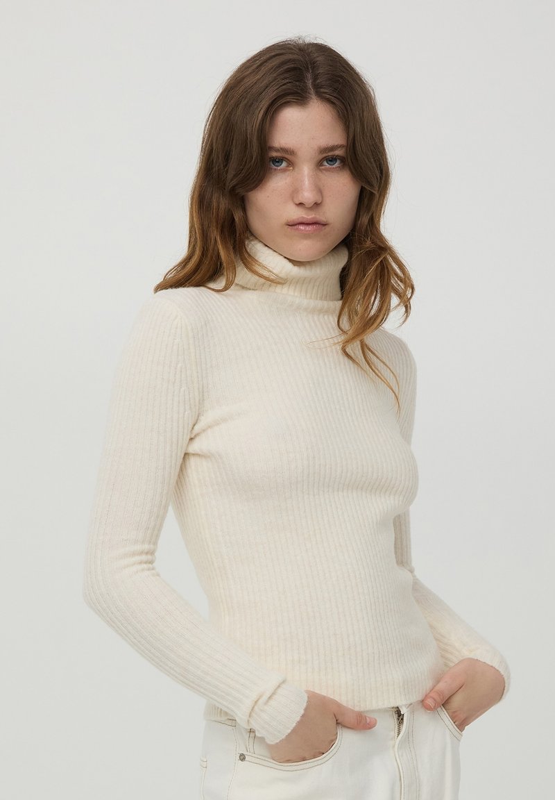 Maglione a collo alto in crema a coste con maniche lunghe e design aderente, realizzato in un materiale morbido e leggero. Presenta un décolleté alto.