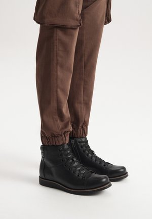 Czarne skórzane botki za kostkę z okrągłym noskiem, sznurowanym zapięciem i zamkiem błyskawicznym. Połączone z brązowymi spodniami z elastycznym mankietem. Gładka tekstura, solidna podeszwa.