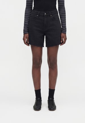 Person iført sorte denimshorts, sorte sokker, sorte sneakers og en langærmet sort og gråstribet top, stående mod en hvid baggrund.