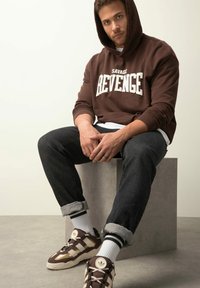 Felpa marrone con testo "SAVAGE REVENGE", jeans scuri e scarpe da ginnastica marroni. Il modello è seduto su un blocco di cemento.