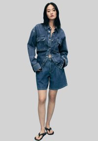 Chemise en denim avec deux poches avant, déboutonnée sur un crop top. Associée à un short en denim assorti. Tongs noires avec des accents cloutés.