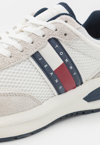 Zapatilla blanca con parte superior de malla, acentos de ante gris y una franjas azul marino con detalles rojos y blancos. Diseño con cordones.