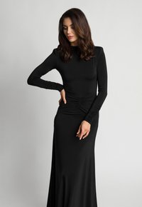 Kobieta o falowanych brązowych włosach ubrana w długą czarną sukienkę z marszczonymi detalami, pozująca z jedną ręką na biodrze i patrząca w dół.