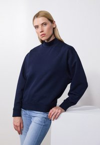 Sudadera navy con cuello alto, puños y dobladillo de canalé. La modelo se apoya en un pedestal blanco, vestida con jeans azul claro.