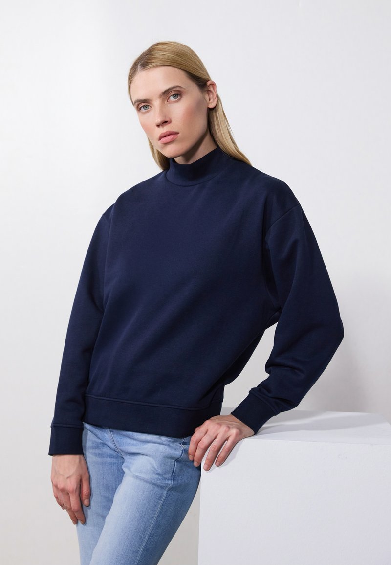 Sudadera navy con cuello alto, puños y dobladillo de canalé. La modelo se apoya en un pedestal blanco, vestida con jeans azul claro.