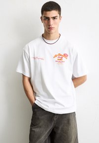 Hvid oversized t-shirt med rød grafik, tekst og motiver på brystet, parret med mørke denimshorts; glat bomuldsstruktur, rund halsudskæring.