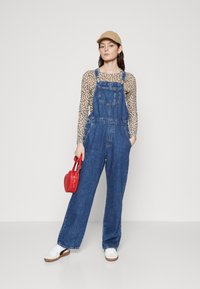 Jeans kombinezoni v srednje modri barvi z širokimi nogami, stranskimi gumbi in sprednjim žepom, v kombinaciji z zgornjim delom v leopardjem vzorcu, belimi supergami in rdečo torbico.