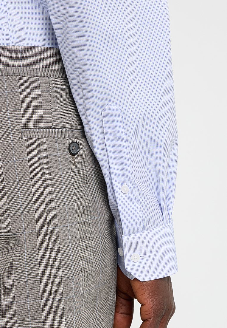 Camisa de vestido de patrón azul claro con puños abotonados, combinada con pantalones de cuadros grises. La camisa presenta pequeños puntos y botones blancos.