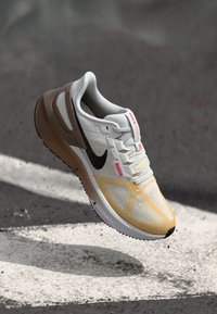 Nike trkačka obuća s bijelim mrežastim gornjim dijelom, žutim detaljima i crnim swooshom. Ima podstavljeni potplat i teksturirane uzorke na tijelu.