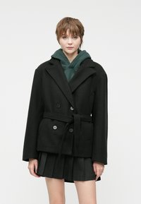 Black double-breasted coat with large lapels, a tie belt, and side pockets, layered over a green hoodie and a pleated black skirt.