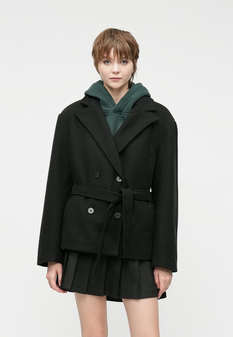 Black double-breasted coat with large lapels, a tie belt, and side pockets, layered over a green hoodie and a pleated black skirt.