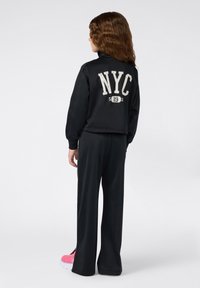 Jong persoon met golvend bruin haar, gekleed in een zwart NYC-jack en bijpassende broek, staande met de rug naar de kijker, met felroze sneakers.