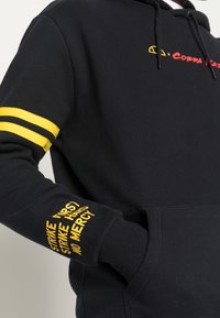 Felpa nera con strisce gialle sulla manica sinistra, con testo giallo sulla manica destra. Materiale morbido con tasca frontale e cappuccio.