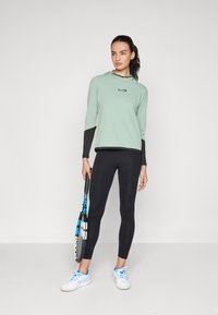 Lysegrønn langermet treningstop med svarte detaljer, parret med svarte leggings og hvite sko. Holder en tennisracket.