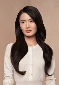 Jeune femme aux longs cheveux ondulés et foncés porte un cardigan blanc boutonné, regardant légèrement sur le côté sur un fond beige.