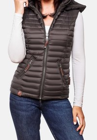 Brun vadderad väst med huva, försedd med dragkedja framtill, två sidofickor och ribbad textur. Bärs över en vit långärmad topp.