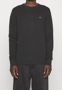 Černý svetr s dlouhým rukávem se strukturovaným vzorem, kulatým výstřihem a žebrovanými manžetami. Malý logo akcent na hrudi.