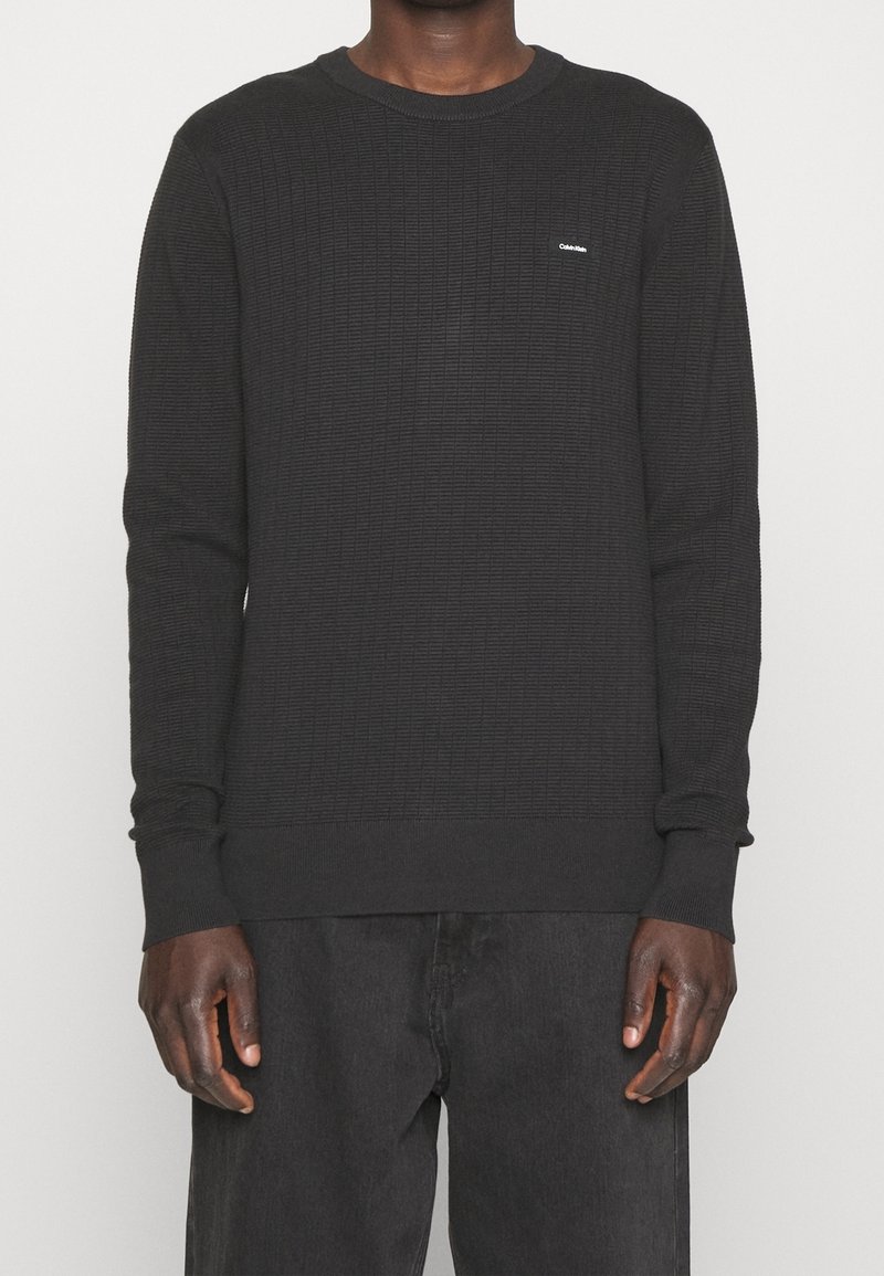 Černý svetr s dlouhým rukávem se strukturovaným vzorem, kulatým výstřihem a žebrovanými manžetami. Malý logo akcent na hrudi.