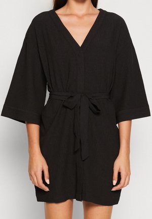 Kvinde iført en sort knælængde kjole med vide ærmer og et bundet bælte i taljen, stående mod en ensartet hvid baggrund.