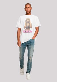Weißes, übergroßes T-Shirt mit einem grafischen Druck einer Person und dem Namen "britney" in Pink, kombiniert mit ausgewaschenen hellblauen Jeans und weißen Sneakers.