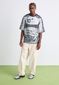 Jovem com cabelo trançado, vestindo uma t-shirt desportiva prateada oversized da Adidas, calças largas em creme e ténis cinzentos, a estar de pé sobre um piso verde.
