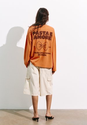 Person mit langen Haaren steht mit dem Gesicht zur Wand, trägt ein oranges Langarmshirt mit Nudelgrafik und beige knielange Cargoshorts.