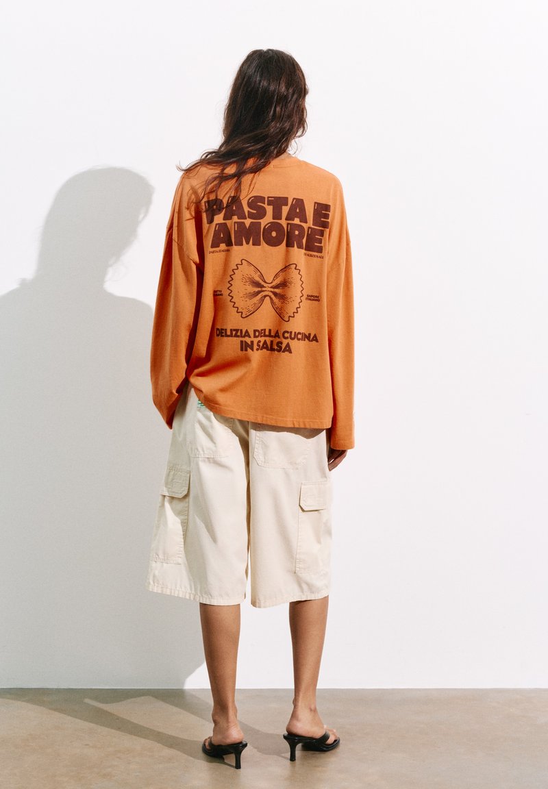 Personne aux cheveux longs se tenant face à un mur, portant un t-shirt orange à manches longues avec un motif de pâtes et un short cargo beige jusqu'aux genoux.