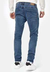 Jean en denim bleu avec une coupe slim, dotés de poches arrière avec des détails de couture. Portés avec des baskets blanches et noires.