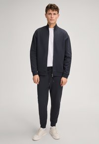 Marineblaues Zip-Up-Jackett mit hohem Kragen, kombiniert mit passender Hose und weißen Sneakers. Aus glattem Stoff gefertigt, mit gerippten Bündchen und Taillenbund.