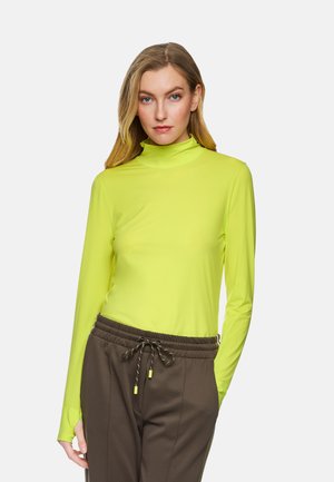 Neon-gelbes Langarm-Turtleneck-Shirt aus leichtem Stoff, kombiniert mit braunen Hosen mit Kordelzug und feiner Struktur.