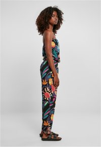Mono con estampado floral en negro con colores vibrantes, top sin tirantes, cintura elástica y piernas ajustadas. Combinado con sandalias negras.