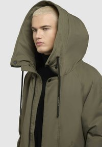 Olivgrön parka med stor huva, framzipper och dragsnören. Tyget har en strukturerad yta och är vattentåligt.