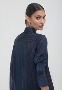Donna con capelli scuri raccolti in un basso chignon, che indossa orecchini a cerchio d'oro e una blusa striata blu navy trasparente, voltandosi verso la spalla contro uno sfondo semplice.