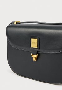 Černá kožená crossbody taška s křivkovým tvarem, zlatými detaily a předním zapínáním. Disponuje texturovaným povrchem a logem jako akcentem.