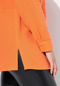 Maglia arancione a maniche lunghe con texture, orlo curvo e spacchi laterali, con polsini abbottonati e vestibilità casual. Abbinata a pantaloni neri.
