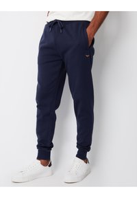 Marineblå sweatpants lavet af blødt stof, med et elastisk bælte og snøre, samt sidelommer; parret med hvide sneakers.