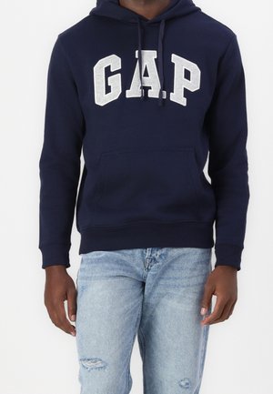 Personne portant un sweat à capuche GAP bleu marine avec logo blanc et un jean déchiré bleu clair, debout devant un fond uni.