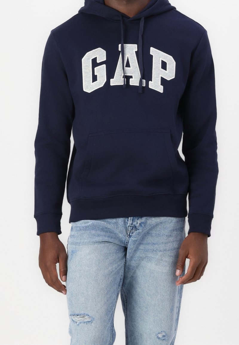 Personne portant un sweat à capuche GAP bleu marine avec logo blanc et un jean déchiré bleu clair, debout devant un fond uni.