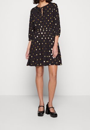 Kobieta ubrana w czarną sukienkę do kolan w złote kropki, z rękawami o długości 3/4 i czarne buty Mary Jane na szerokim obcasie.