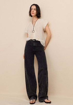 Giovane donna con capelli corti e scuri che indossa una camicetta crema larga, jeans neri a gamba larga con motivi floreali, infradito nere e una collana con ciondolo.