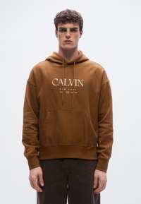 Brun hoodie av mjukt material med frampicka och dragsnören. Har vit "CALVIN KLEIN"-logotyp. Enkel design.