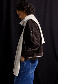 Brun bomberjakke med sidelommer og lomme på ærmerne, parret med et lys gråt tørklæde draperet over skulderen. Bæret med blå jeans.