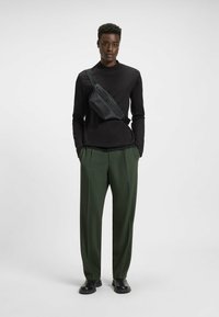 Schwarzes Langarmshirt kombiniert mit grünen plissierten Hosen. Eine schwarze Umhängetasche wird quer über die Brust getragen. Glatte Texturen und eine elegante Silhouette.